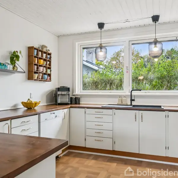 Køkken med hvide skabe og mørke bordplader, kaffemaskine og køkkenredskaber på hylder. To store vinduer viser grønne buske udenfor. To hængelamper lyser rummet op.