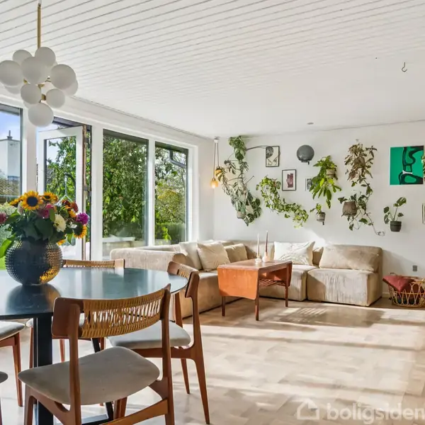 Spisestuebordet står med blomsterbuket, under moderne loftslampe, i lys stue med store vinduer. Sofa med puder, vægudsmykning og TV på stativ danner hyggelig hjørne.