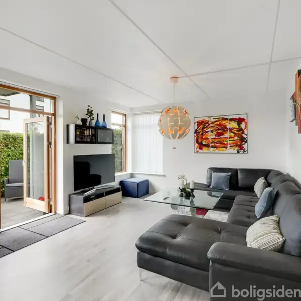 Grå hjørnesofa står i en stue med hvidt gulv. Et glasbord foran, farverig kunst hænger på væggen. Store vinduer og døre åbner ud til terrasse med grønne omgivelser.
