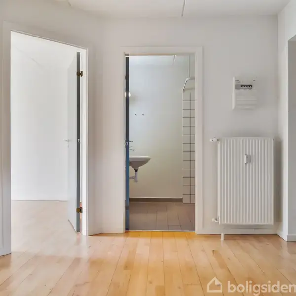 En lys gang med trægulv har fire åbne døre førende til forskellige rum, herunder et badeværelse med synligt håndvask. En radiator og elektrisk kontrolpanel er monteret på væggen.