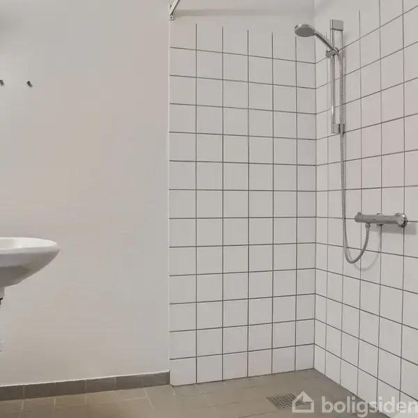 Håndvask og bruser i hvidt, flisebelagt badeværelse med lysegrå gulve. Bruseren har et vægfæstet armatur, og vasken hænger ved siden af. Enkel og minimalistisk indretning.