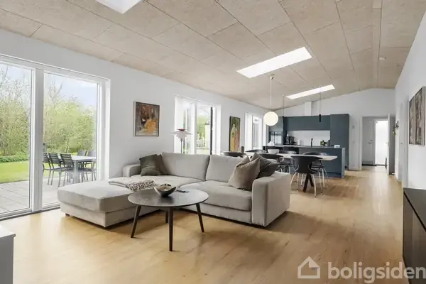 Sofa står i en moderne stue med parketgulv, ved siden af et rundt bord. Rummet har store vinduer mod en terrasse, og køkkenet ses i baggrunden.