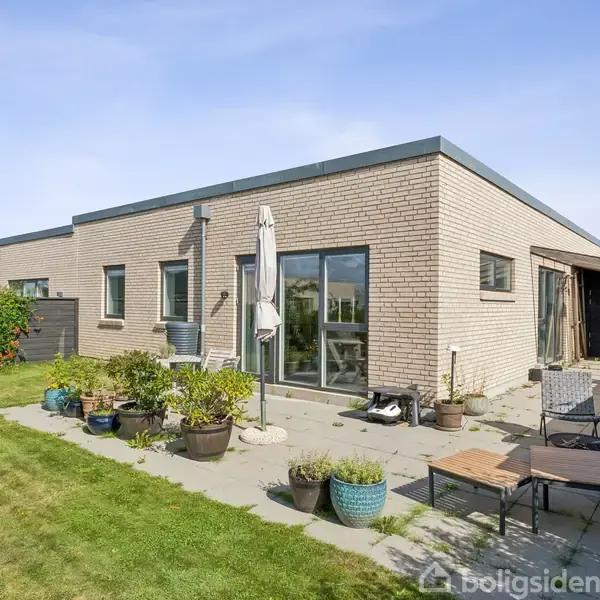 En moderne, fladtaget murstensbygning står i en have. Den har store vinduer ud mod en terrasse med møbler og plantekrukker. Græsplænen omgiver terrassen, og himlen er klar.