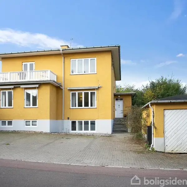 Et gult hus med hvide vinduer og en balkon står ved siden af en garage og indkørsel. Omgivet af grønne buske og træer, på en stille gade.