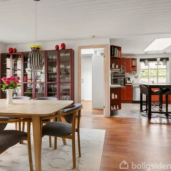 Spisebord med stole står i et lyst køkken-alrum. Baggrund med røde skabe, køkkenø, og store vinduer. En vase med blomster er midt på bordet.
