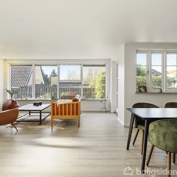 Stue med møbler såsom en lænestol, sofa og spisebord. Rummet er lyst med store vinduer, der slipper naturligt lys ind, og en glasdør fører ud til en terrasse.