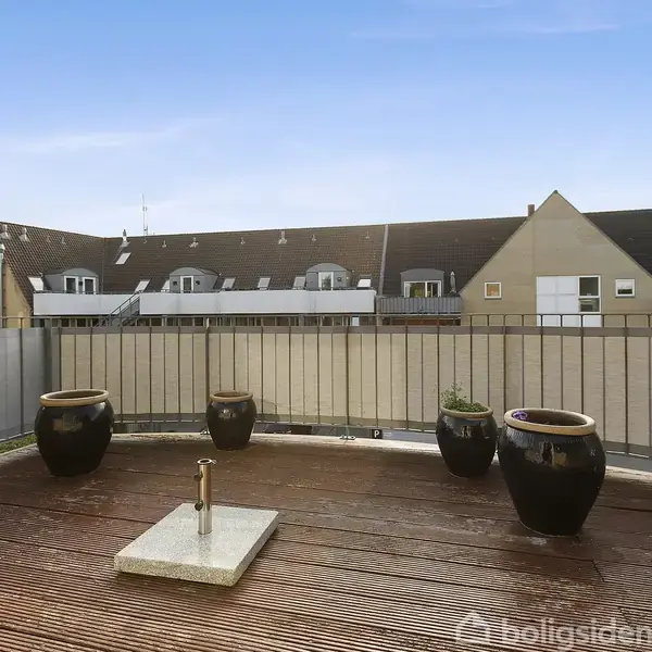 En træterrasse med tre sorte krukker står stille. Den er omgivet af et gelænder og en murstensvæg. I baggrunden er der en række af bygninger med stejle tage.
