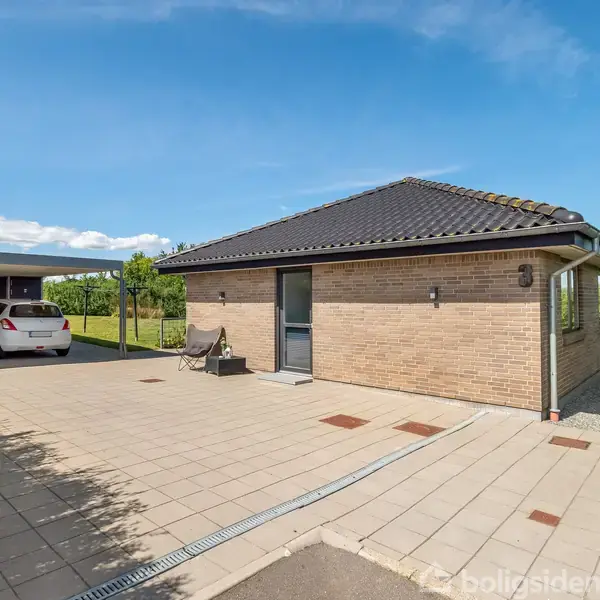 Et enkeltplanshus i mursten med sort tag står midt i en flisebelagt indkørsel. Til venstre er en carport med to parkerede biler, omgivet af grønt landskab under en blå himmel.