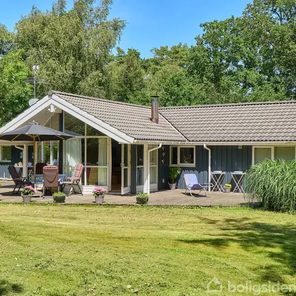 Sommerhus med gråt tag og blå vægge, står på en græsplæne omgivet af skov. En terrasse med parasol, stole og planter foran.