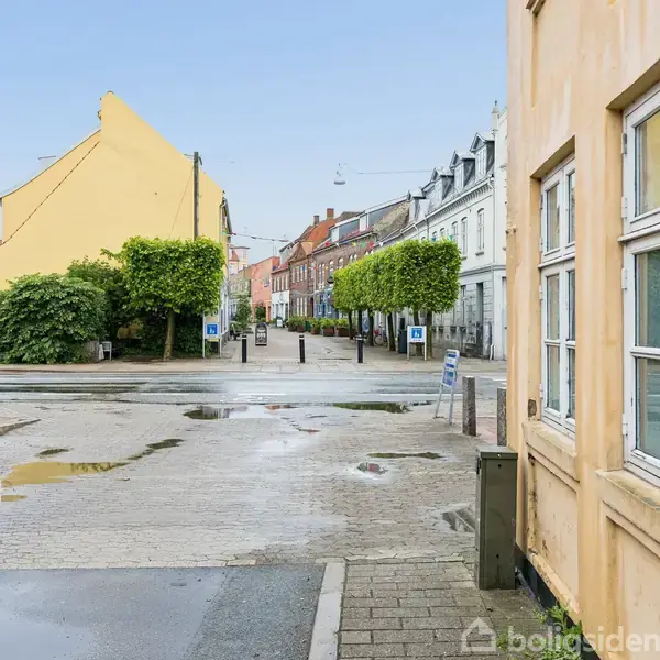 Gade med farverige bygninger langs en våd vej. Træer står i række til højre. Rundt skilt på væggen viser nummer 36, og teksten er "Porsborgsvej 36".
