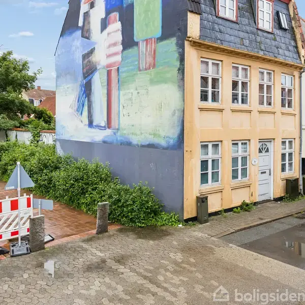 Bygning med stor vægmaleri af is i boligområde, parkering foran med hvid varevogn, og vejafspærring til venstre. Tæt på grønne buske og træer.