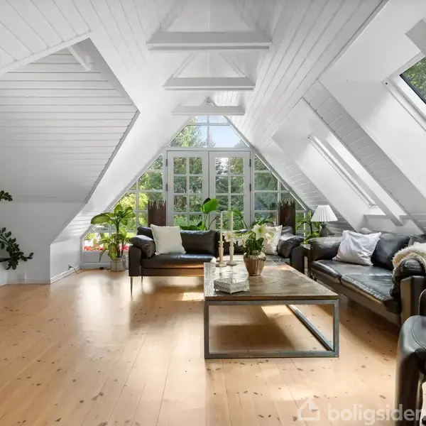Et lysekronebelyst stueområde med skråt hvidt loft, store vinduespartier, to lædersofaer og et træbord. Rummet er dekorativt med grønne planter og har trægulve.