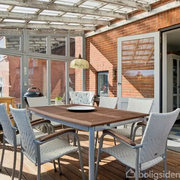 En overdækket terrasse med et træbord omgivet af seks stole. Terrassen har glaspartier og en murstensvæg med en dør til en indendørs spiseplads. Grill og planter står langs væggen.