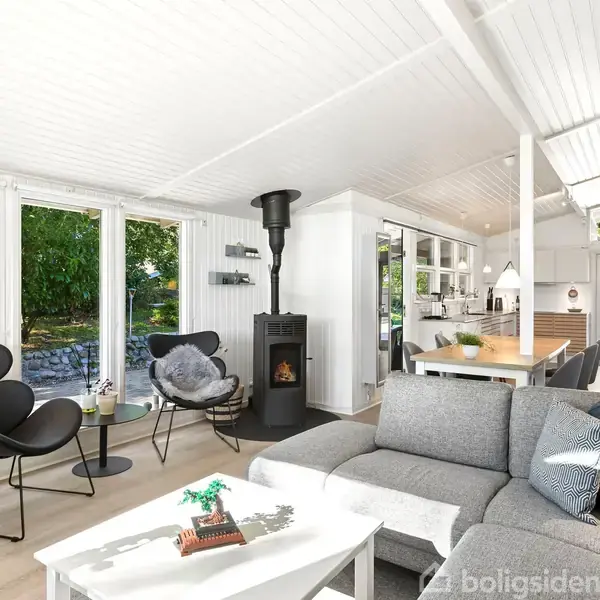 Grå sofa står i et lyst, moderne stueområde med et lille bord foran. Brændeovn brænder til højre, og store vinduer viser en have med grønne træer udenfor.