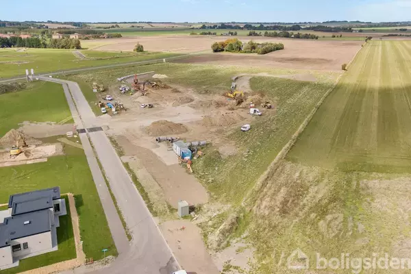 Byggeplads med flere gravemaskiner arbejder på et jordområde omgivet af åbne marker og veje. I forgrunden ses et moderne hus ved en asfalteret vej.