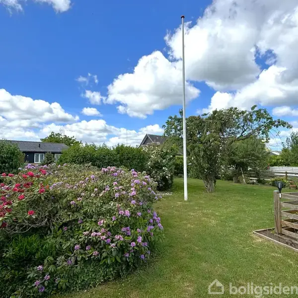 Flagstang står stille i en have, omgivet af frodigt grønt græs og blomstrende buske. Baggrunden viser en blå himmel med skyer og et hus.