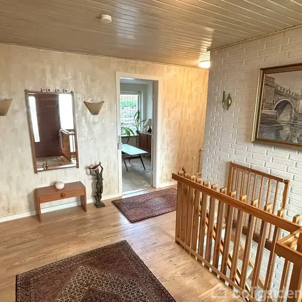 Gang med trægulv og trappe; væg med maleri af bro. Borde med spejl og dekorative genstande. Dør åbner til andet rum med planter. Jakker hænger til venstre.