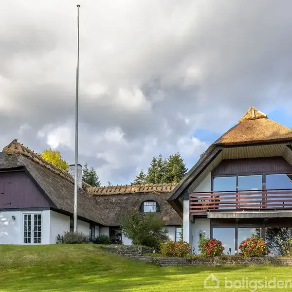 Stråtækt hus står stille i et frodigt haveområde med græs og blomster. Husets facade har store vinduer og balkon, mens himlen er dækket af tunge, grå skyer.