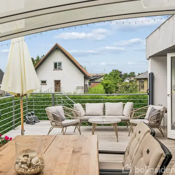 En overdækket terrasse viser et spisebord med stole, parasoller og en siddegruppe. Terrassen har udsigt til en boligkvarter med huse og grønne haver i baggrunden.