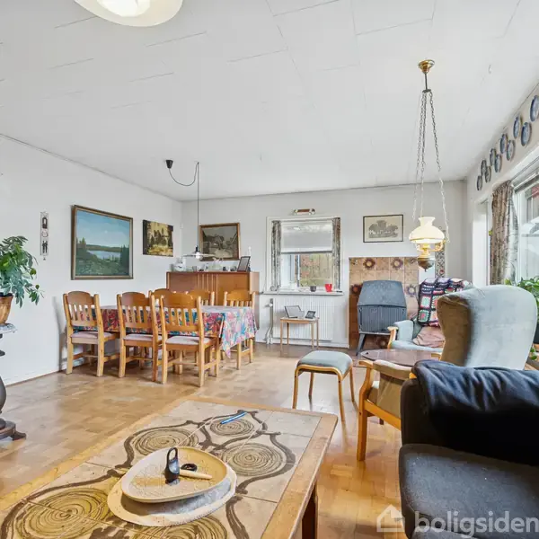 Stue med træmøbler inkluderer en sofagruppe og spisebord med stole. Væggene er dekoreret med malerier og tallerkener. Store vinduer til højre giver udsigt og lysindfald.