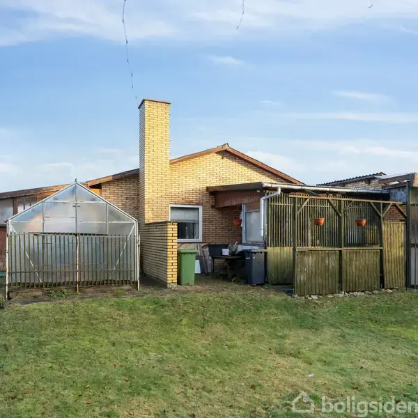 Et enfamiliehus i gule mursten står i en have med græsplæne. En glaskonstruktion og skure ses til venstre og højre for huset under en klar blå himmel.