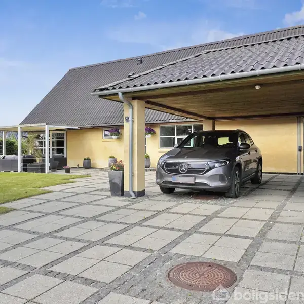 Bil parkeret under carport ved moderne gult murstenshus med sort tag. Terrasse med havemøbler ses til venstre, omgivet af græsplæne og enkelte træer.