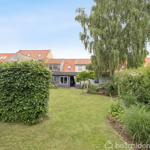 Et rækkehus med rødt tegltag og grå facade står i en have med grøn græsplæne, buske og træer. Området er omgivet af andre ens rækkehuse med tilsvarende tage.