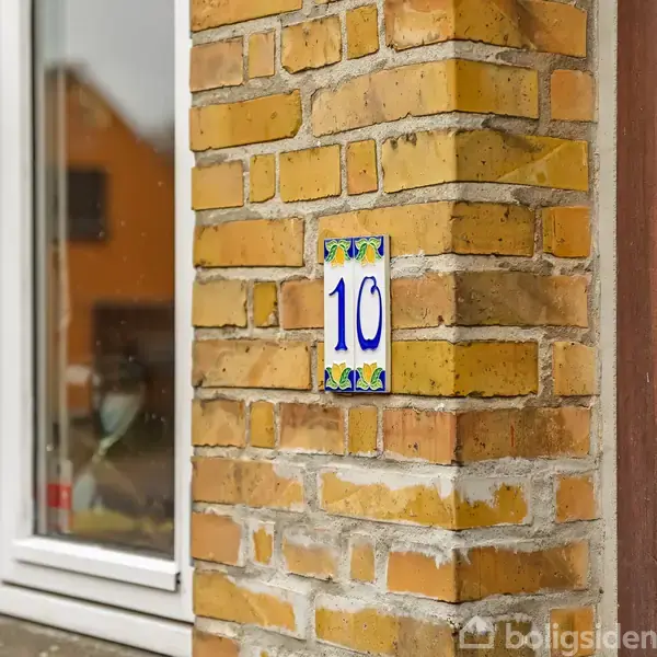 Et husnummer "10" er placeret på en murstensvæg ved siden af en trædør. I nærheden ses et vindue med planter indendørs, og i baggrunden skimtes en orange bygning.