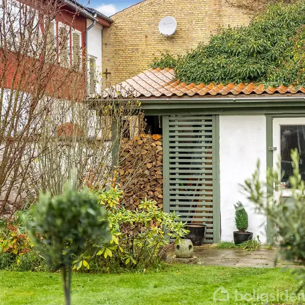 Et lille havehus med hvid facade og rødt tegltag står i en frodig have. Træer og buske omgiver huset, og brændestabler er opbevaret langs siden.