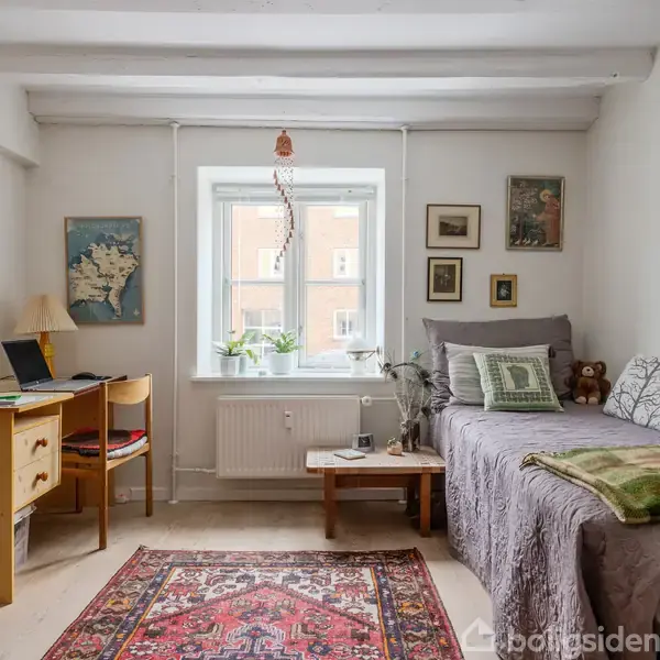 En lille, hyggelig stue med gråt tæppe på en sofa. Et skrivebord med lampe og laptop står til venstre. Væggen har forskellige billeder, og et tæppe ligger på gulvet.