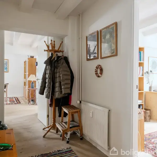 Frakke hænger på en træstativ ved siden af en stige i en lys gang med malerier på væggen og døråbninger til værelser med tæpper og møbler.