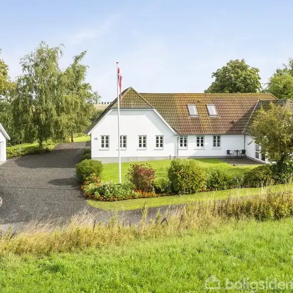 En hvid villa med brun tag beliggende på en velplejet, grøn græsplæne. Ejendommen har en indkørsel af grus, en garage og en flagstang med et dannebrog.