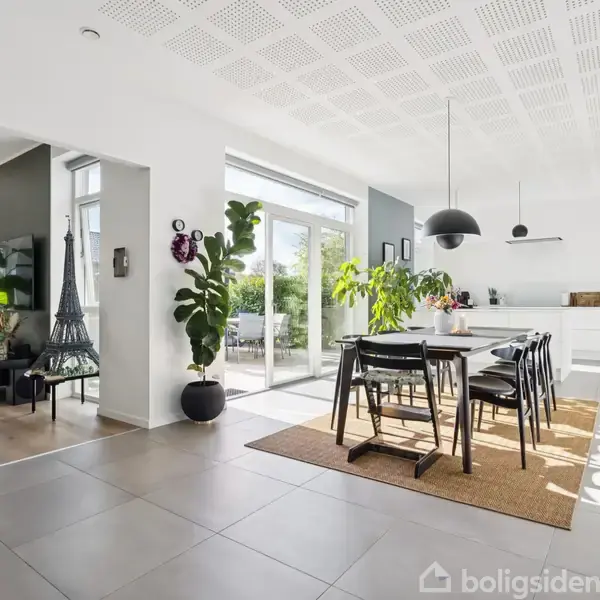 Spisebord med sorte stole står på et tæppe midt i et lyst køkken-alrum med store vinduer. Rummet har moderne indretning med grønne planter og minimalistisk belysning.