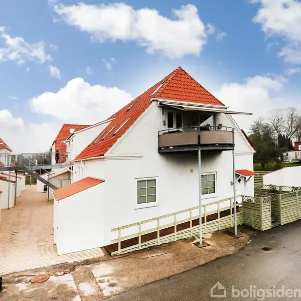 Et hvidt hus med rødt tegltag har en lille, rund balkon. Huset ligger i et beboelsesområde med flere lignende bygninger under en blå himmel med skyer.