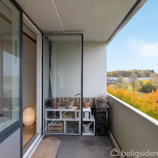 En altan med planter i potter står stille ved en glaskant. Bygninger og efterårstræer omgiver altanen i et boligområde med klart himmel.