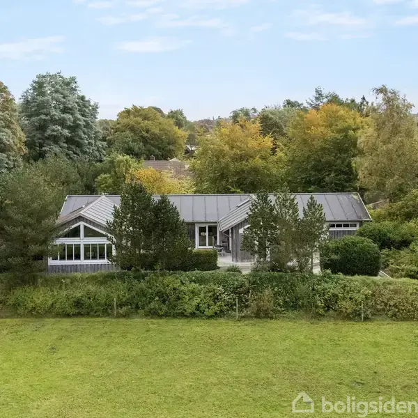 Et hus med gråt tag og store vinduer omgivet af frodig vegetation og træer. Foran huset er en stor grøn græsplæne. Baggrunden viser en skov og blå himmel.