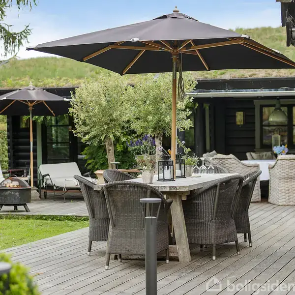 Et træbord med kurvestole står på en terrasse med store sorte parasoller. Der er et sort hus i baggrunden omgivet af grønne planter og en grill.