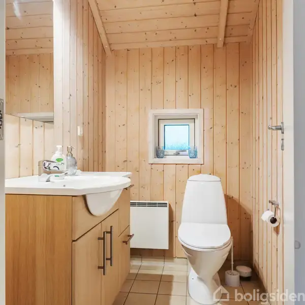 Toilet og vask i træbeklædt badeværelse. Toiletrulle på væggen, skab under vasken, og lille vindue nær loftet. Flasker på vasken. Væggene er lysebrune og rummet ser hyggeligt ud.