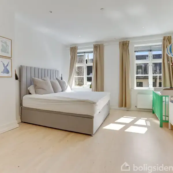 Sengen står i et lyst soveværelse med trælaminatgulv, puder, og sengegavl, ved siden af vinduer med beige gardiner. Der er en gul stol, grøn skrivebord og lyse vægge.