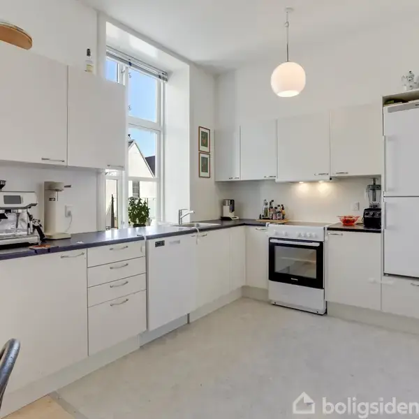 Køkken med hvide skabe og en sort bordplade, indeholder kaffemaskine og komfur. Lys hænger fra loftet, og store vinduer tillader dagslys at strømme ind.