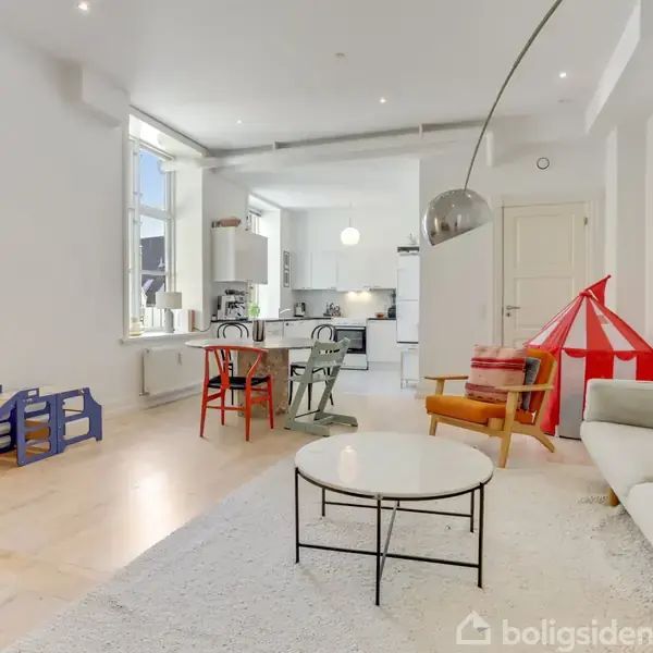 Stue med sofa til højre, rundt sofabord og stor gulvlampe. Venstre side har børnerutsjebane. Baggrund viser et køkken med spisebord og stole. Hvidt og lyst interiør.