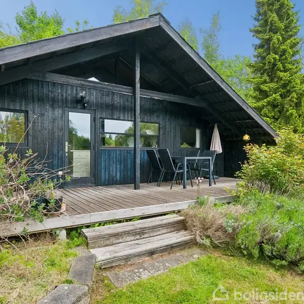 Et sortmalet træhus med terrasse indeholder et udendørs bord og stole. Huset er omgivet af grønne træer og buske i et naturligt miljø.