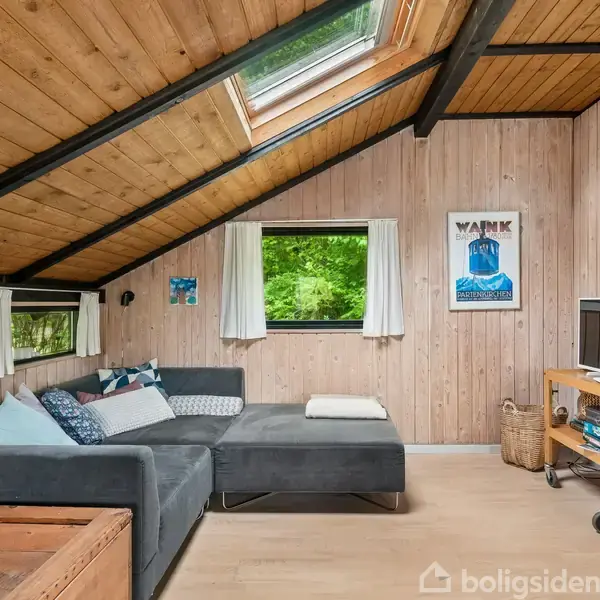 Stue med stor grå sofa står under skråt loft med træpaneler og ovenlysvindue. Et lille TV er placeret på en rullereol, og væggen har et "NAKKE" plakat.