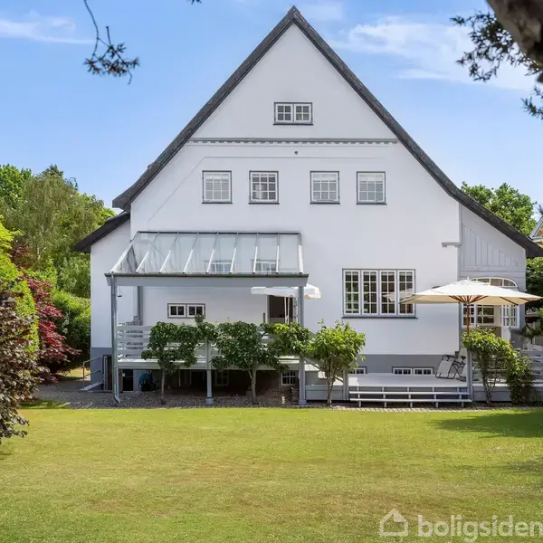 Hvid villa med skrånende tag, solskærm og glasoverdækket terrasse. Grønne planter og plæne omgiver huset i et velplejet, solrigt haveområde.