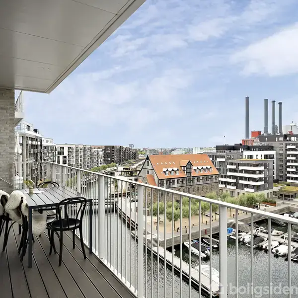 En altan med et bord og fire stole, der er dækket af tæpper, tilbyder udsigt over en moderne bydel med kanaler og både. Lejlighedsbygninger omgiver området under en blå himmel.