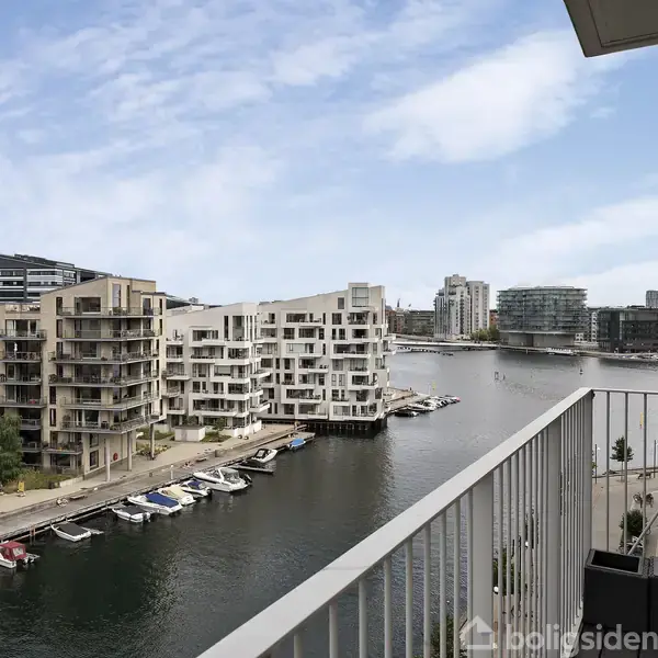 En altan med udsigt til en kanal, hvor flere både ligger til kaj. Omgivet af moderne, lyse beboelsesejendomme og en delvis overskyet himmel.