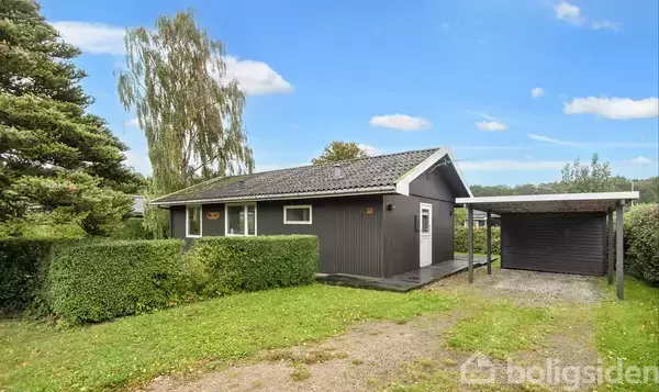 Et gråmalet sommerhus står stille, omgivet af en grøn have med træer og en carport til højre, mod en klar blå himmel med få skyer.