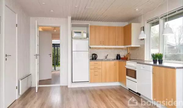 Køkken i lyst træ med køleskab, komfur, kaffemaskine og planter står i et lyst rum med trægulv og vinduer, som giver udsigt til udendørs grønne arealer.