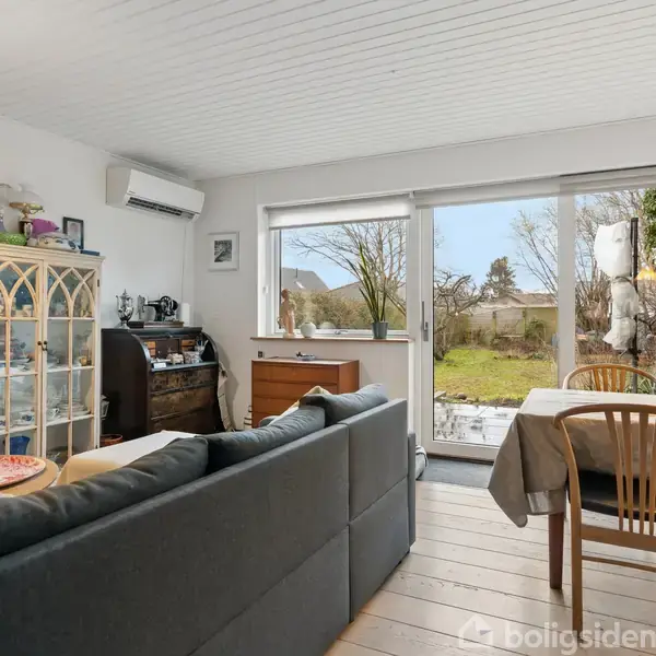 En stue med en sofa, et spisebord og stole. Rummet har store vinduer, der vender mod en have. Der er en bogreol i hjørnet og en aircondition på væggen.