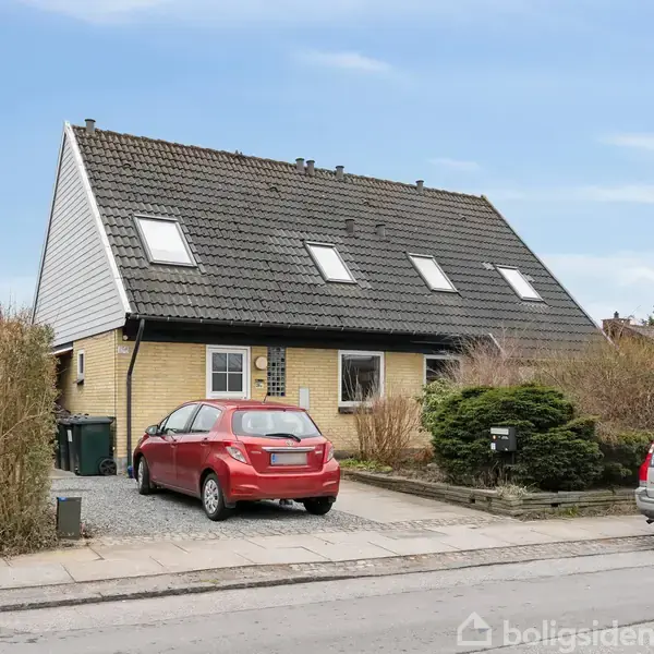 En murstensvilla med skråt tag står på en villavej. En rød bil er parkeret i indkørslen, og nogle buske omgiver huset. To biler holder på vejen.